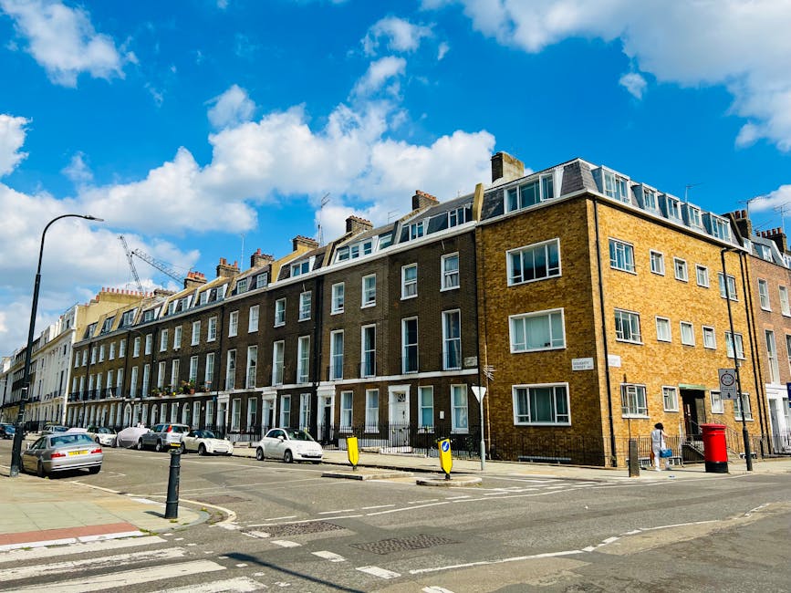 A multi-storey residential building on Kings Road viewed from the street, featuring a brick façade with numerous white-framed windows and a flat roof with dormer windows. The ground level has entry doors and small front gardens, with cars parked along the curbside. The sidewalk is paved, and a red post box and streetlamp are visible near the building. Bright daylight illuminates the scene against a backdrop of a blue sky with scattered white clouds. The image showcases urban residential architecture typical of Chelsea, London, as seen in the context of Kings Road flat cleaning rates and property maintenance services, with Cleaner Chelsea providing surface cleaning and deep cleaning solutions in the area.