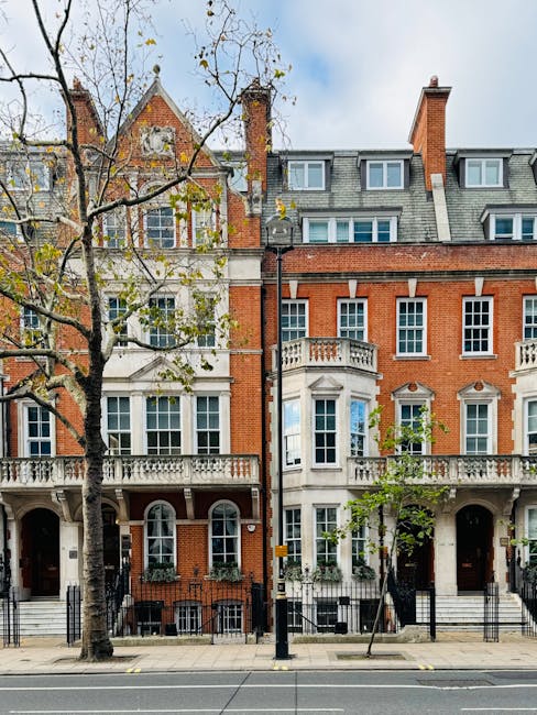 Exterior view of a row of historic residential buildings on Kings Road in Chelsea, featuring red brick facades, white decorative stone window sills, and balconies. The buildings have large sash windows, some with flower boxes, and are topped with slate roofs and chimneys. In front, a sidewalk is lined with leafless trees and black metal railings, with a lamppost standing near the curb. The overall scene shows a tidy, well-maintained streetscape under a partly cloudy sky, illustrating the charming architecture typical of the SW3 area, as highlighted by Cleaner Chelsea's expertise in surface cleaning and deep cleaning services for residential properties.