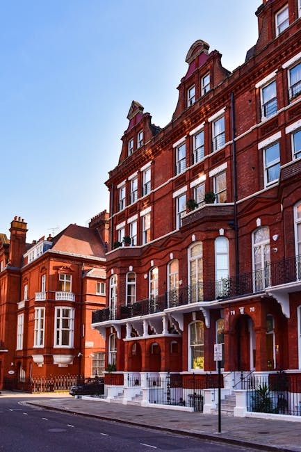 A multi-storey residential building on Kings Road viewed from the street, featuring a brick façade with numerous white-framed windows and a flat roof with dormer windows. The ground level has entry doors and small front gardens, with cars parked along the curbside. The sidewalk is paved, and a red post box and streetlamp are visible near the building. Bright daylight illuminates the scene against a backdrop of a blue sky with scattered white clouds. The image showcases urban residential architecture typical of Chelsea, London, as seen in the context of Kings Road flat cleaning rates and property maintenance services, with Cleaner Chelsea providing surface cleaning and deep cleaning solutions in the area.